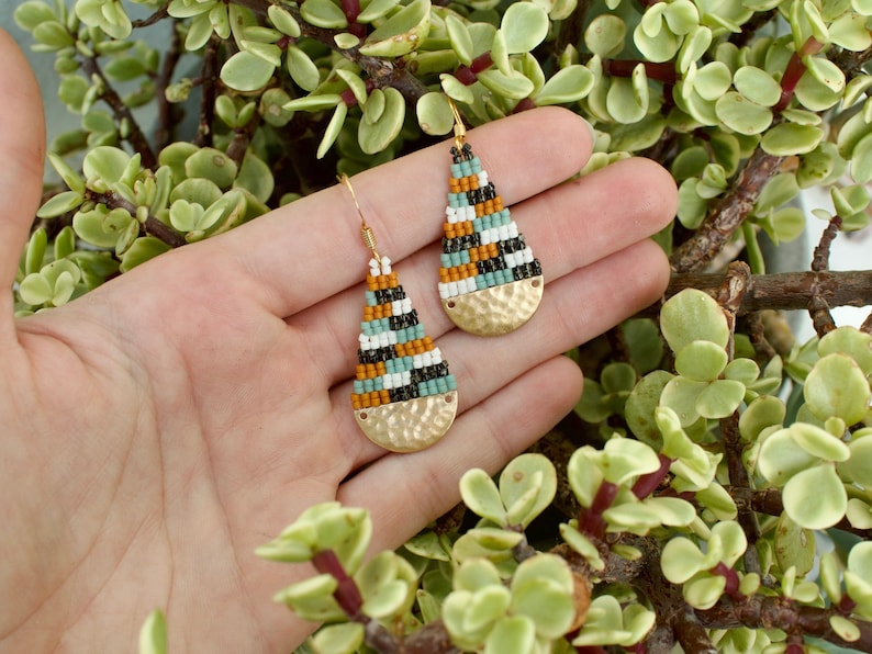 May include: A pair of teardrop-shaped earrings with a hammered gold finish. The earrings are made with black, white, brown, and teal beads.