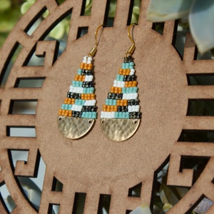 May include: A pair of gold tone earrings with a geometric pattern of white, black, orange, and blue beads. The earrings have a hammered metal disc at the bottom.