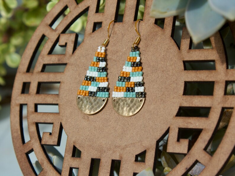 May include: A pair of gold tone earrings with a geometric pattern of white, black, orange, and blue beads. The earrings have a hammered metal disc at the bottom.