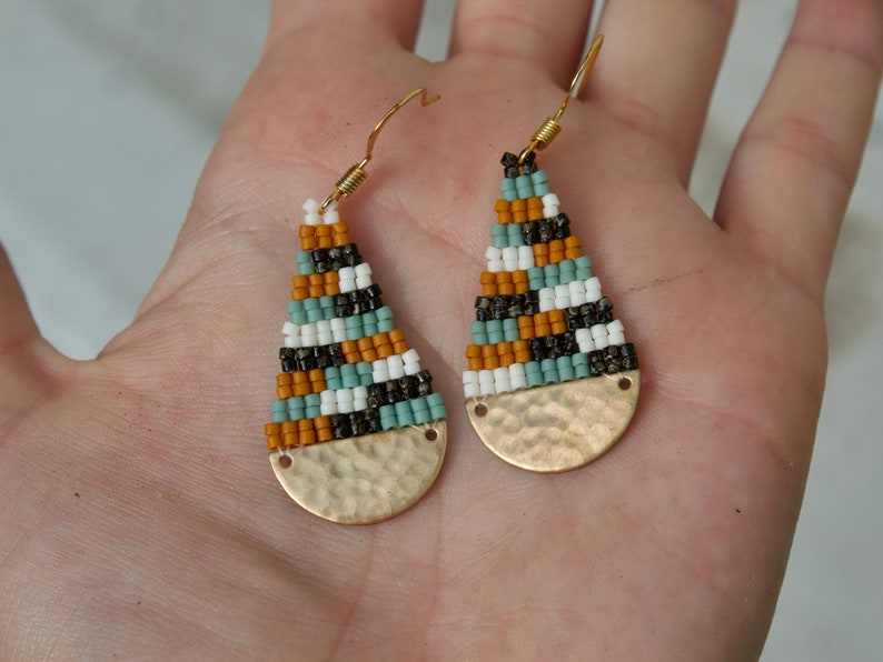 May include: A pair of gold teardrop earrings with a hammered metal bottom. The earrings are beaded with a geometric pattern of black, white, orange, and teal beads.
