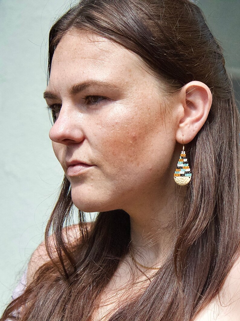 May include: A woman with long brown hair wearing a pair of teardrop-shaped earrings with a beaded design in shades of blue, orange, and white.