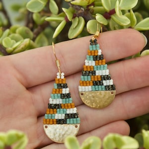 May include: A pair of teardrop-shaped earrings with a geometric pattern of black, white, orange, and teal beads. The earrings have a gold-toned hammered metal base.