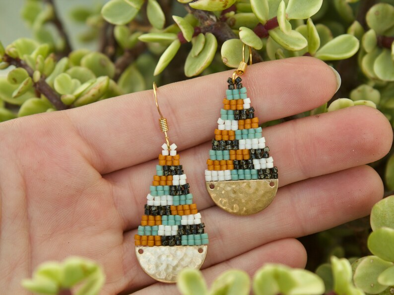 May include: A pair of teardrop-shaped earrings with a geometric pattern of black, white, orange, and teal beads. The earrings have a gold-toned hammered metal base.