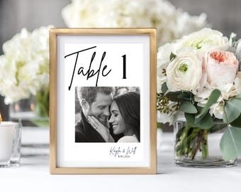 May include: A framed table number card with a gold frame. The card features the word "Table 1" in a handwritten font, a black and white photo, and names and a date at the bottom. The background includes flowers and a lit candle.