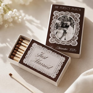 Può includere: Due scatole di fiammiferi rettangolari con copertine marrone scuro e bordi bianchi in stile pizzo. Una scatola è aperta, mostrando fiammiferi in legno, con il testo Just Married. L'altra presenta una foto in bianco e nero di una coppia, The Perfect Match, 05 November 2026.