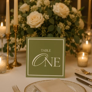 Puede incluir: Una mesa de boda con un centro de mesa floral, velas y una tarjeta con el número de mesa. La tarjeta es de color verde oliva con la palabra "TABLE" encima de la palabra "ONE" en escritura blanca. Un plato con borde dorado con una servilleta doblada y cubiertos están en primer plano.