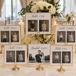 May include: Gold framed table number signs with black and white photos of children and a couple. The signs are displayed on a white tablecloth with floral arrangements and candles. The signs read "Table One" through "Table Five" and "Bridal Party."