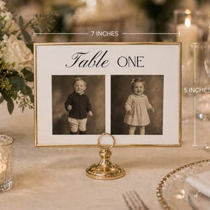 Puede incluir: Una tarjeta con n&uacute;mero de mesa con marco dorado, 17.78 cm de ancho y 12.7 cm de alto, con el texto "Table ONE" sobre dos retratos vintage de ni&ntilde;os. La tarjeta se sujeta con un soporte dorado decorativo, adecuado para eventos.