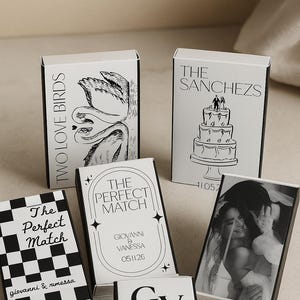 Może przedstawiać: Kolekcja białych pudełek z zapałkami z czarnym tekstem i grafiką. Projekty obejmują ciasto, parę, szachownicę oraz napisy "Two Love Birds" i "The Perfect Match". Niektóre pudełka zawierają imiona i daty.