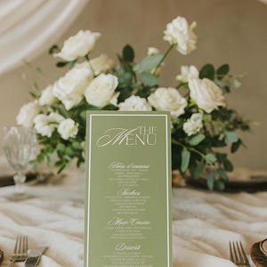 May include: A green menu card with white text, featuring a floral arrangement of white roses. The menu card has a white border and lists various food courses. Silverware and a glass are visible.