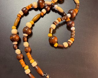 May include: A beaded eyeglass chain featuring a variety of wooden and plastic beads in shades of brown, tan, yellow, and cream. The strand includes round, cylindrical, and textured beads, finished with black rubber loops at both ends for attachment.