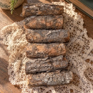 May include: A stack of six cylindrical wooden logs with rough, textured brown bark. The logs are arranged horizontally in a single column, showcasing natural wood grain and dark, weathered bark patterns on their exterior surfaces.