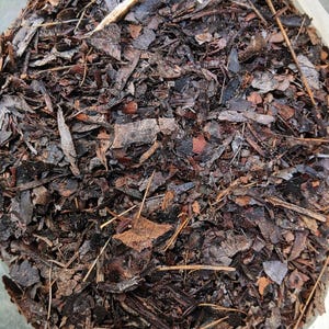 May include: A close-up view of a pile of organic mulch. The mulch consists of various sizes of brown and dark brown wood chips, bark, and dried leaves. Some light-colored straw-like pieces are also visible.