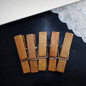 May include: Five wooden clothespins with silver metal springs are arranged in a row. The clothespins are a warm brown color. A white paper and a white lace fabric with blue accents are in the background.
