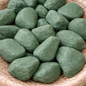 May include: A collection of smooth, green decorative stones fills a woven basket. The stones vary in size and shape, with a matte finish. The basket is made of natural, light-colored woven material. The overall aesthetic is earthy and natural.