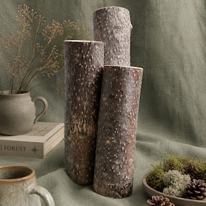 May include: Three cylindrical wooden vases with textured bark, varying in height, stand on a green cloth. A small ceramic mug and a vase with dried plants are in the background. A wooden bowl with moss and pinecones is also present.