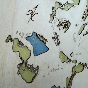 May include: A layered wooden map of a golf course featuring laser-cut details. The map displays numbered holes 12 through 18, with green fairways, a blue water hazard at hole 14, and a small label for the clubhouse. A north arrow is visible.