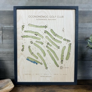 May include: A framed wooden map of the Oconomowoc Golf Club in Oconomowoc, Wisconsin. The light wood surface features green-tinted golf course fairways, a blue water hazard, and black text detailing the club name, architect, and course statistics.