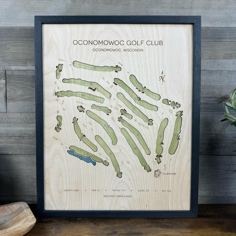 May include: A framed wooden map of the Oconomowoc Golf Club in Oconomowoc, Wisconsin. The light wood surface features green-tinted golf course fairways, a blue water hazard, and black text detailing the club name, architect, and course statistics.