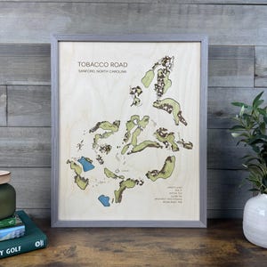 May include: A framed wooden map of the Tobacco Road golf course in Sanford, North Carolina. The light-colored wood surface features green fairways, brown sand traps, and blue water hazards, with text detailing course statistics and history.