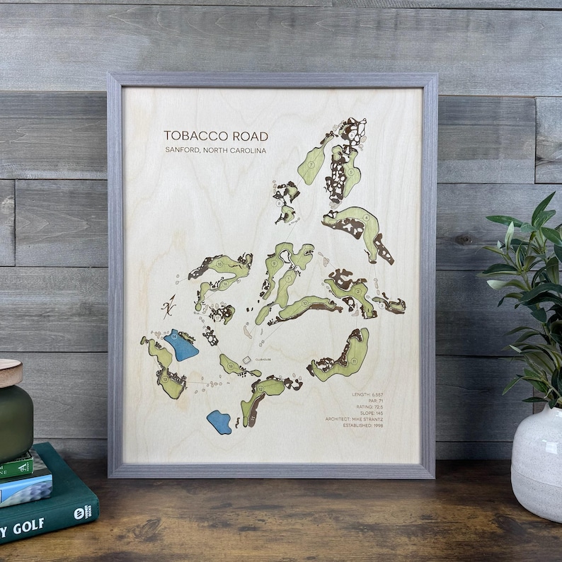 May include: A framed wooden map of the Tobacco Road golf course in Sanford, North Carolina. The light-colored wood surface features green fairways, brown sand traps, and blue water hazards, with text detailing course statistics and history.