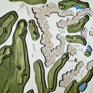 May include: A detailed wooden golf course map with green fairways, beige sand traps, and blue water features. The map includes hole numbers and the word "HOME" with a house icon. The design is laser-cut and layered.