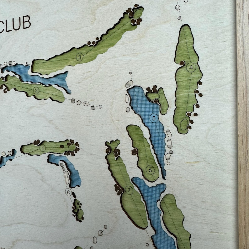 May include: A laser-cut wooden golf course map featuring light green fairways and blue water hazards. The map includes numbered holes from one to eight, with dark brown sand traps and a light wood frame border. The text CLUB is visible in the corner.