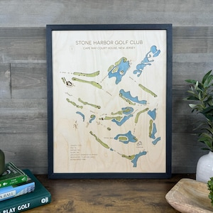 May include: A framed wooden map of the Stone Harbor Golf Club in Cape May Court House, New Jersey. The map features light wood grain, blue water hazards, and green fairways. Text includes course details, architect, and establishment year.