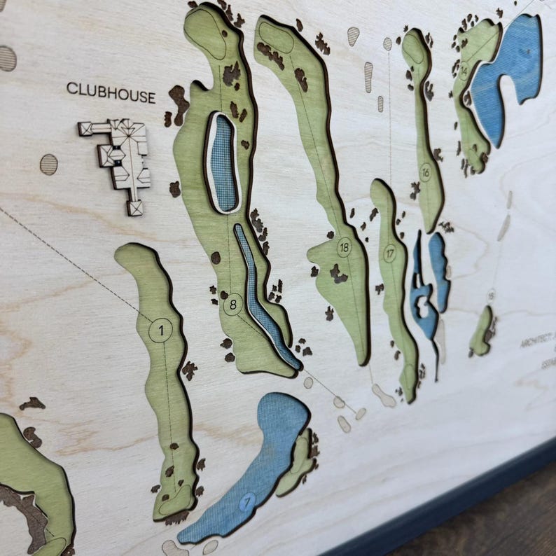 May include: A layered wooden golf course map featuring light green fairways, blue water hazards, and dark brown sand traps. The map includes engraved hole numbers, a clubhouse icon, and the text CLUBHOUSE, all set against a natural wood grain background.