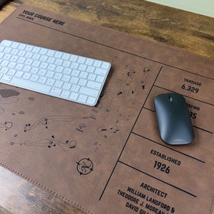 May include: A brown leather desk mat featuring a printed golf course layout, the text YOUR COURSE HERE, CITY, STATE, YARDAGE 6,329, RATING 73.5, ESTABLISHED 1926, and ARCHITECT WILLIAM LANGFORD & THEODORE J. MOREAU & DAVID GILL, with a white keyboard and mouse.