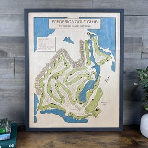 May include: A framed wooden map of the Frederica Golf Club in St. Simons Island, Georgia. The map features light wood tones for land, blue for water, and green for fairways. Text includes course statistics, the club name, and a compass rose.