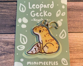 May include: A leopard gecko keyring with a gold-coloured clasp and a light green backing card. The keyring features a cartoon gecko with yellow and brown spots. The card has the words "Leopard Gecko - Keychain-" and the brand name "minimeeples".