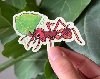 Puede incluir: Una pegatina troquelada que muestra una hormiga roja estilizada cargando una hoja de color verde brillante. La hormiga tiene un cuerpo rojo oscuro con reflejos naranjas, grandes ojos negros y mandíbulas prominentes. Pequeñas chispas verdes rodean la cabeza.
