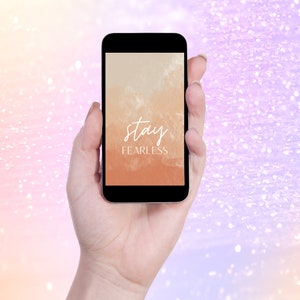 Op de afbeelding: Een zwarte smartphone toont de motiverende quote "stay fearless" in witte letters tegen een aquarel-achtige achtergrond van oranje en beige tinten. De telefoon wordt in een hand gehouden tegen een zachte, glinsterende achtergrond.