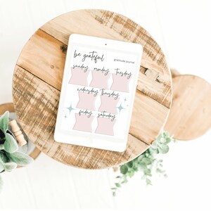 May include: A digital tablet displaying a pink and white printable gratitude journal with the text "be grateful" and "gratitude journal". The journal is divided into seven sections, one for each day of the week, with the days of the week labelled in cursive.