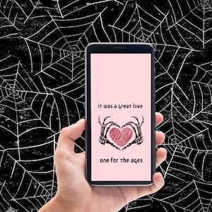 Könnte beinhalten: Ein schwarzes Smartphone mit einem rosa Bildschirm, auf dem der Text "it was a great love one for the ages" zu sehen ist. Der Text ist von einer schwarz-weißen Illustration von zwei Skeletthänden umgeben, die ein rotes Herz halten.
