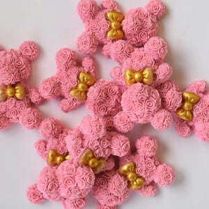May include: A group of pink rose teddy bears with gold bows. The bears are made of small pink roses and have a cute, whimsical look.