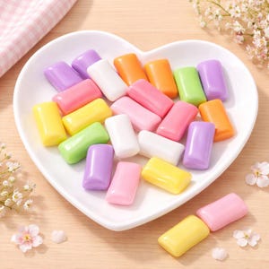 May include: A heart-shaped white dish filled with colorful chewing gum. The gum pieces are rectangular and come in various colors, including pink, purple, orange, green, yellow, and white. The arrangement is on a light wooden surface.