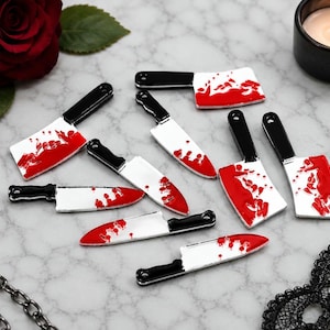 May include: A collection of small metal charms shaped like kitchen knives and meat cleavers. Each charm features a silver-colored blade with red enamel splatters resembling blood and a black enamel handle with a small hole at the end for hanging.