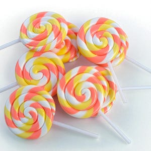 May include: A collection of colorful spiral lollipops. Each lollipop features a swirl design with alternating stripes of white, yellow, and peach. The lollipops are on white sticks, and are arranged on a white surface.