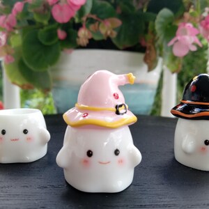 May include: Three white ceramic ghost figurines with smiling faces and rosy cheeks. One ghost wears a pink witch hat with yellow trim, another wears a black hat with orange trim, and the third is a small planter. The figurines are on a dark surface.