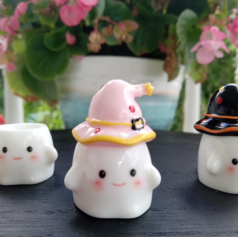 May include: Three white ceramic ghost figurines with smiling faces and rosy cheeks. One ghost wears a pink witch hat with yellow trim, another wears a black hat with orange trim, and the third is a small planter. The figurines are on a dark surface.