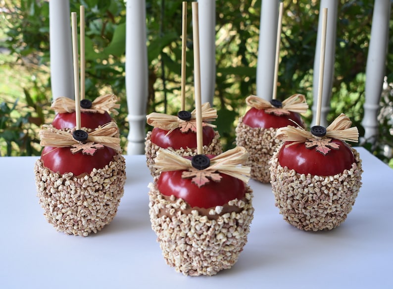 May include: Five red caramel apples decorated with brown and tan straw, a brown button, and a small red leaf. The apples are on wooden sticks and are arranged on a white surface.