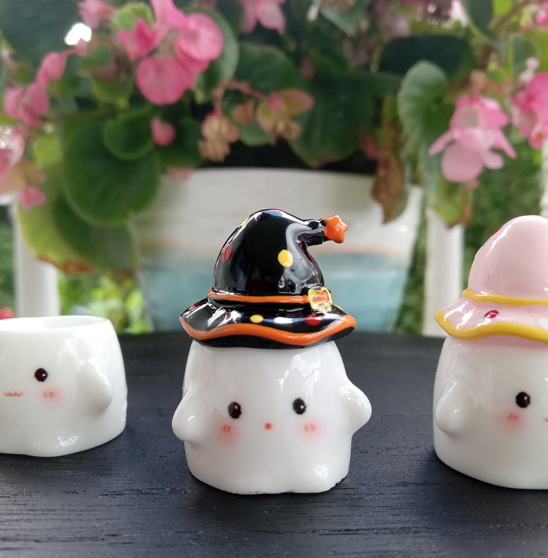 May include: Three white ceramic figurines with cute faces. One wears a black witch hat with orange and yellow accents. Another wears a pink hat with a yellow brim. The third figurine is a small, open container. The background features pink flowers.