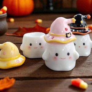 May include: A collection of Halloween-themed ceramic figurines. The figurines are white with smiling faces and are wearing witch hats in yellow, pink, and black. Candy corn and fall leaves are scattered around the figurines on a wooden surface.