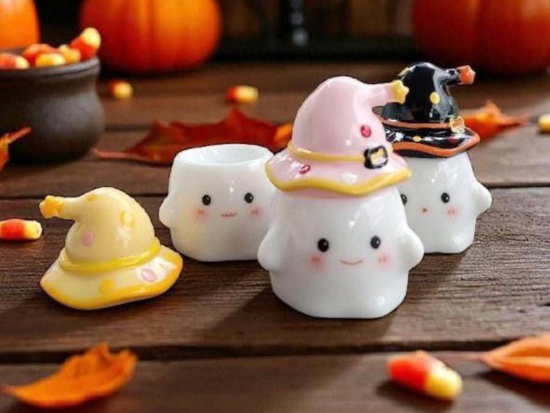 May include: A collection of Halloween-themed ceramic figurines. The figurines are white with smiling faces and are wearing witch hats in yellow, pink, and black. Candy corn and fall leaves are scattered around the figurines on a wooden surface.