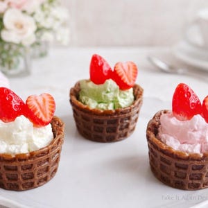 May include: Three miniature desserts in waffle cups, each topped with a different colored cream and a fresh strawberry. The desserts are arranged on a white plate, with a blurred background of flowers and a teacup.