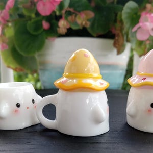 May include: White ceramic mushroom-shaped mugs and a small planter with smiling faces and blush cheeks. One mug has a yellow hat with orange trim, another has a pink hat with red accents. The planter has a similar design. Decorative and whimsical.