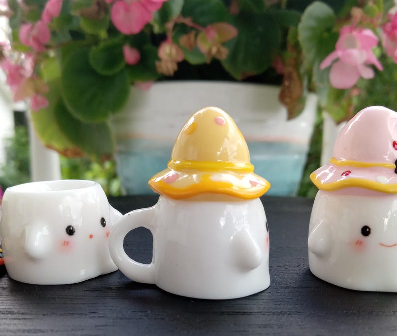 May include: White ceramic mushroom-shaped mugs and a small planter with smiling faces and blush cheeks. One mug has a yellow hat with orange trim, another has a pink hat with red accents. The planter has a similar design. Decorative and whimsical.