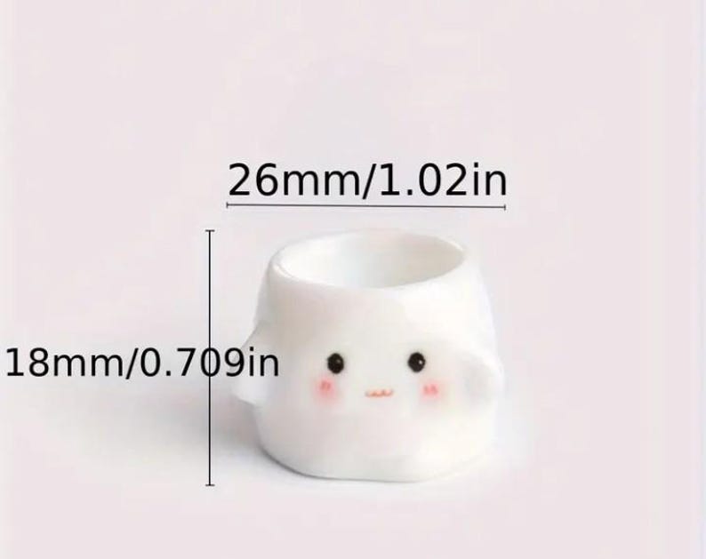May include: A small, white ceramic planter with a cute face design. The planter is 1.02 inches wide and 0.709 inches tall. It features black eyes, rosy cheeks, and a small mouth. The planter has small, rounded ears.